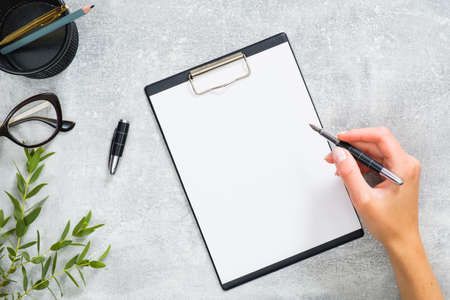 Woman hand holding pen and write a message in blank paper clipboard. Feminine home office desk with stationery, glasses, branch with green leaves on concrete stone background. Business concept.の写真素材