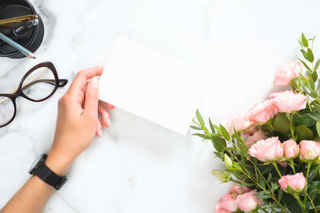 Woman hand holding blank paper card mockup over modern home office desk workspace with stationery, rose flowers bouquet, glasses, Flat lay, top view.の写真素材