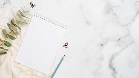 White marble background with blank paper notepad, eucalyptus, scarf and office supplies. Flat lay, top view, copy space. Home office desk table concept.の写真素材
