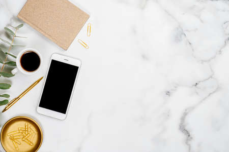 White marble office desk table with smartphone screen mockup, gold paper notebook, eucalyptus leaf, pen, cup of coffee and stationery. Flat lay, top view, copy space.の写真素材
