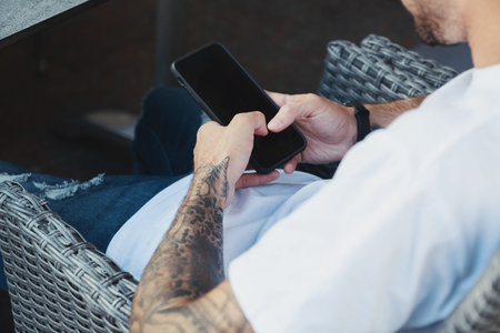 Tattoo hands holding mobile phone. Handsome man dressed white t-shirt sitting in coffee shop and...