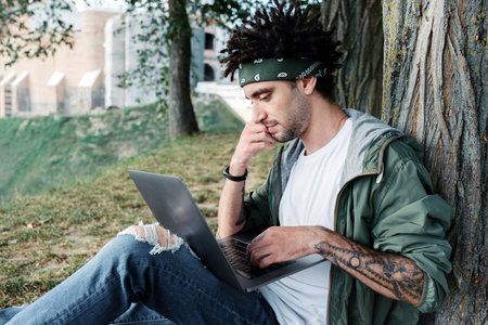 Young millennial student with dreadlocks sitting on grass and using portable laptop computer outdoor...