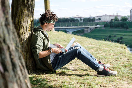 Young millennial student with dreadlocks sitting on grass and using portable laptop computer outdoor in public park. Freelancer using notebook for distance job, remote working, e-learningの写真素材