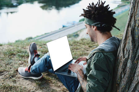 Rear view of young hippie freelancer working outdoor, sitting on the grass, using laptop with white screen mockup, typing on keyboard. DIgital nomad work remote on new startup projectの写真素材