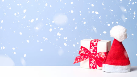 Little white gift box with red ribbon bow and Santa Claus hat in snow on blue background. Christmas banner mockup, New Year greeting card template.