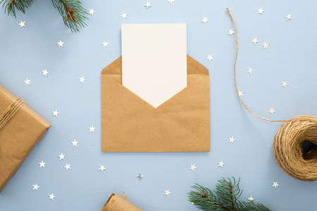Empty blank card in kraft paper envelope on pastel blue background decorated with confetti star, gift box, wrapping paper and fir tree branches. Christmas, New Year, winter holiday invitation mockup.の写真素材