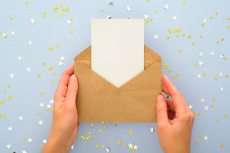 Female's hands holding kraft paper envelope letter with blank white card mockup over pastel blue background with golden confetti stars. Christmas, New Year, winter holidays and birthday concept.の写真素材