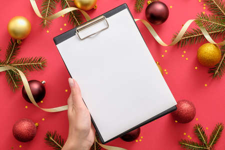 Hand holding clipboard mockup with blank paper over Christmas background with Christmas decorations, balls, gold ribbon, fir tree branches on red background. Checklist, New Year goals conceptの写真素材