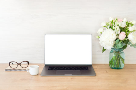 Office desk table with laptop computer with blank screen mockup and flowers bouquet, coffee cup, glasses, notepad on wooden surface. Minimal style feminine workplace concept.の写真素材