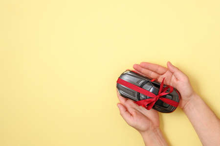 New car gift concept. Toy car with red ribbon in human hands on yellow background. Top view. Auto dealership and rental, raffle prize, lottery drawing, car buying or selling.の写真素材