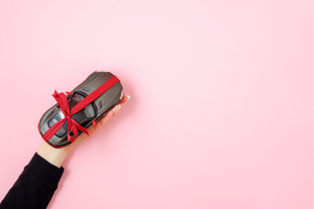 Woman's hand holding toy car model tied with a red ribbon and bow as gift on pink background. Car...