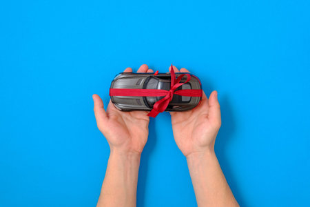 New car gift concept. Toy car model tied with a red ribbon and bow in human hands on blue...