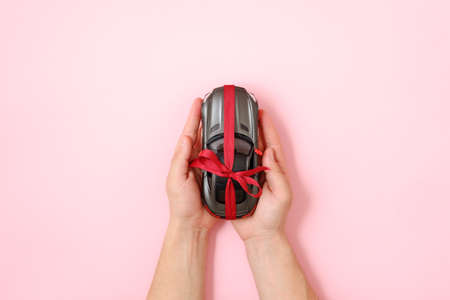 Human hands holding toy car model tied with a red ribbon and bow on pink background. Car insurance services, car for sale, special offer, draw a prize concept. Flat lay composition. Top view.の写真素材
