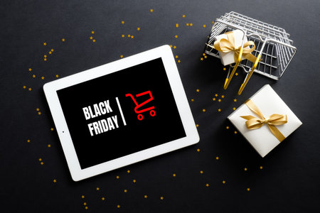 Black Friday sale concept. Tablet with sign "Black Friday" on screen, shopping basket, gift box, confetti over black background. Flat lay, top view, overhead.の写真素材