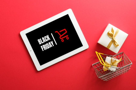 Black Friday concept. Tablet with sign Black friday on screen, shopping cart, gift box over red...