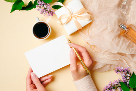 Womens hand holding golden pen and write message on blank paper card on beige feminine background with stylish accessories, perfume bottle, silk scarf, gift box, coffee cup. Greeting card, invitation.の写真素材