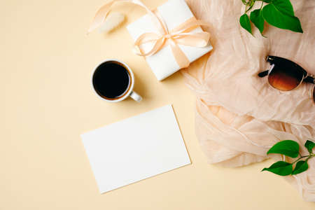 Women workspace with paper card, coffee cup, green leaves, gift box, glasses on beige background. Feminine desktop. Wedding invitation concept. Flat lay, top view.の写真素材