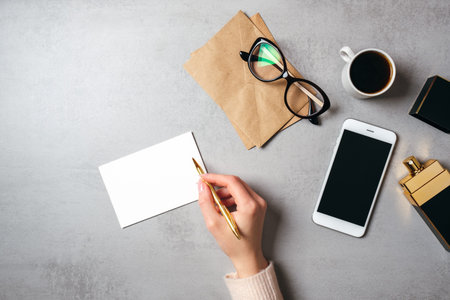 Flat lay modern minimal home workspace with hand of woman writing text on blank paper card, glasses,...