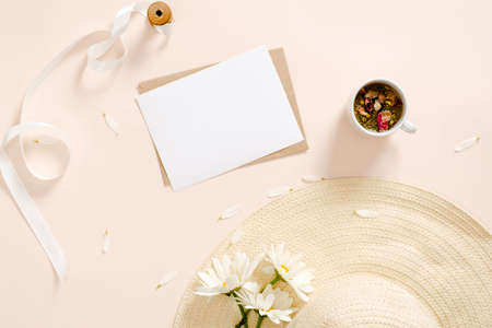 Flatlay composition with blank paper card mockup, envelope straw hat, daisy flowers, tea cup, ribbon on pastel pink background. Flat lay, top view, copy space. Summer fashion, feminine workspace.の写真素材