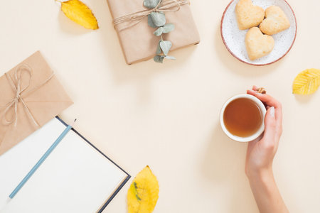 Woman hand holding cup of tea over autumn background with fallen leaves, book, cookies, gift box....