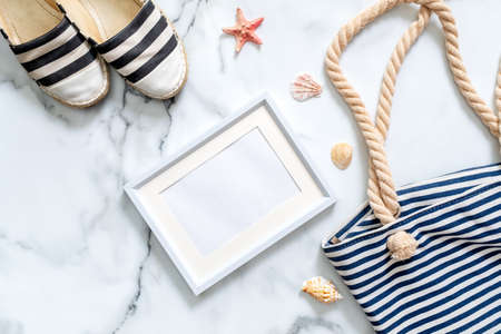 Travel composition on a marble background. Women's desk with striped sandals, beach bag, seashells and blank picture frame. Lifestyle of traveler, beauty blogger, digital nomad, hipster.の写真素材