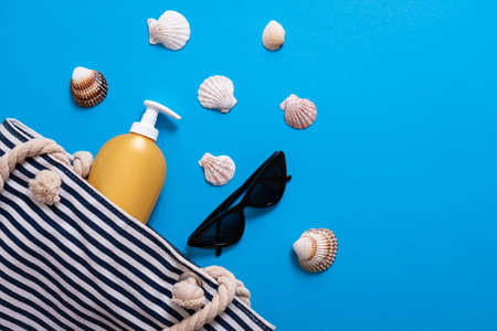 Summer composition. Stripped beach bag with suntan cream, seashell, sunglasses. Summer holiday concept, sunscreen cosmetic and accessories on blue sea background.の写真素材