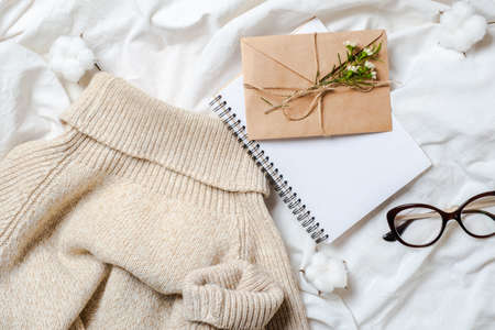 Romance flat lay composition with blank paper notepad, craft paper envelope with flowers, woolen sweater, glasses, cotton on girl's bed. Top view festive feminine accessories. Christmas letter conceptの写真素材