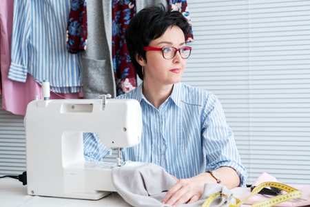 Fashionable seamstress in eyeglasses working at tailor shop with electrical industrial sewing...