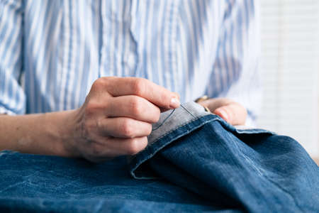 Tailor sew the hem of jeans in workshop, close up view on hands with sewing needleの写真素材