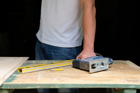 Hands of human holding electric jigsaw lying on workbench with spirit level and yellow pen. DIY...