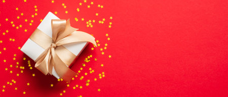 White gift box with golden ribbon bow on red background with confetti stars. Christmas present,...