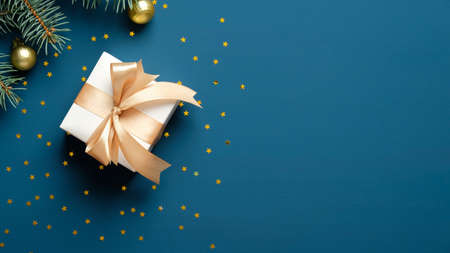 White gift box with golden ribbon bow on blue background with confetti and fir branches. Christmas greeting card template, banner mockup. Flat lay, top view, copy space.の写真素材
