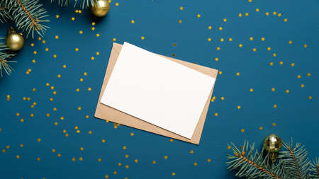 Blank card and craft paper envelope on blue background decorated with confetti and fir branches. Holiday invitation mockup.の写真素材
