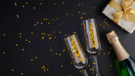 Champagne bottle, glasses, gift box, golden confetti on black background. Christmas or birthday celebration concept. Flat lay, top view.の写真素材