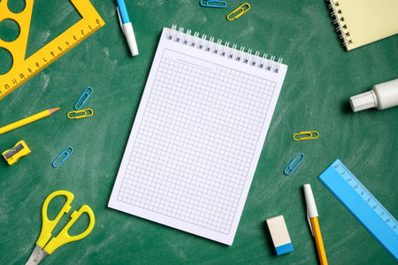 Blank squared notebook and school stationery on green chalkboard background. Flat lay, top view,...