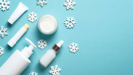 Winter skin care concept. Set of cosmetic products and snowflakes on blue background. Lipstick, tube, moisturizer cream jar, serum dropper bottle, pump bottle. Flat lay, top view.の写真素材