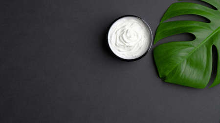 Jar with cosmetic cream and tropical monstera leaf on black background. Natural organic beauty product for body, hand, face skin treatment. Flat lay, top view. Skincare concept.の写真素材
