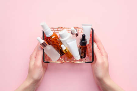 Female hands holding shopping basket with cosmetics on pink background. Beauty products sale concept.の写真素材