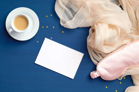 Happy morning concept. Flat lay composition with cup of coffee, sleep eye mask, blanket and empty paper card on dark blue backgroundの写真素材
