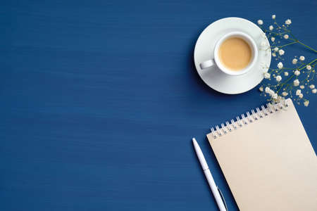 Feminine workspace with cup of coffee, paper notepad and flowers on classic blue background. Flat lay, top view, copy space. Cozy home office desk, hygge, happy morning conceptの写真素材