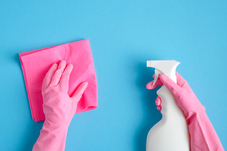 Female hands in pink gloves holding cleaning spray bottle and sanitary napkin on blue background....