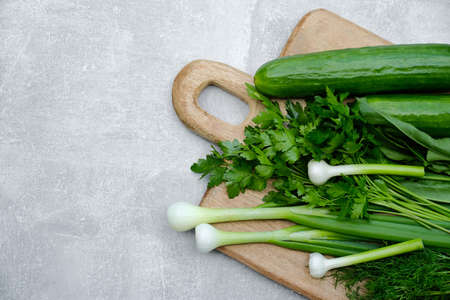 Wooden cutting board with fresh green onion, cucumbers, parsley, dill on stone background. Top view.の写真素材