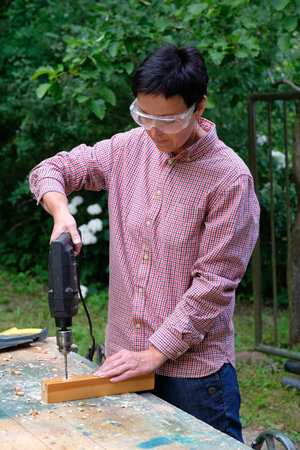 Craftswoman with safety glasses drilling a hole with an electric drill machine in wooden plank....