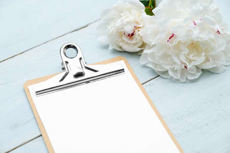 Clipboard blank sheet of paper and peony flowers on blue wooden table. Flat lay, top view. Wedding planner, checklist, to do list concept.の写真素材