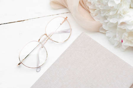 Feminine workspace with glasses, paper notebook and peonies on white wooden table. Elegant flat lay composition, rustic style.の写真素材