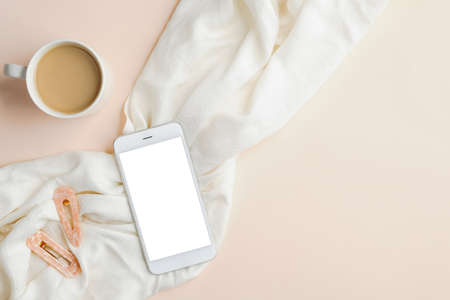 Flat lay cup of coffee, smartphone with blank screen mockup, white blanket and female accessories on beige table. Top view feminine workspace, home office desk.の写真素材