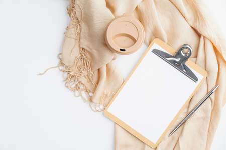 Cozy home office desk with beige blanket, cup of coffee, clipboard with empty paper note on white background. Flat lay, top view. Hygge or nordic style feminine workspace.の写真素材