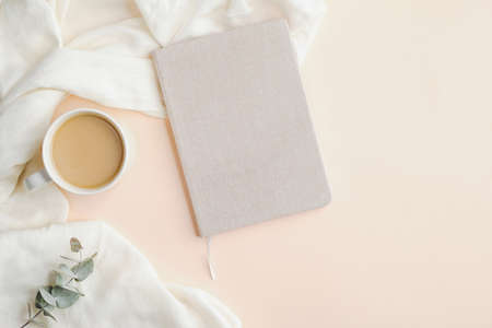Home office desk with paper notebook, cup of coffee, plaid, eucalyptus leaf on beige background. Modern feminine workspace, minimalist style.の写真素材