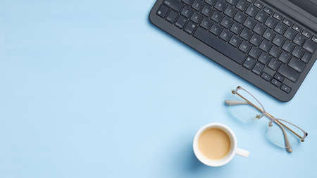 Office desk table with laptop computer keyboard, glasses, coffee cup on blue background. Top view modern desktop, feminine workspace with copy space. Flat lay business conceptの写真素材