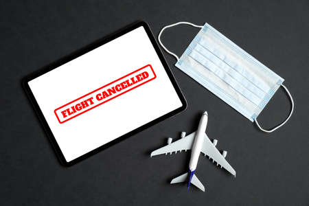 Flight canceled due to coronavirus disease (COVID-19) concept. Face mask, tablet with sign "Flight canceled" on the screen and plane model on black background. Top view.の写真素材